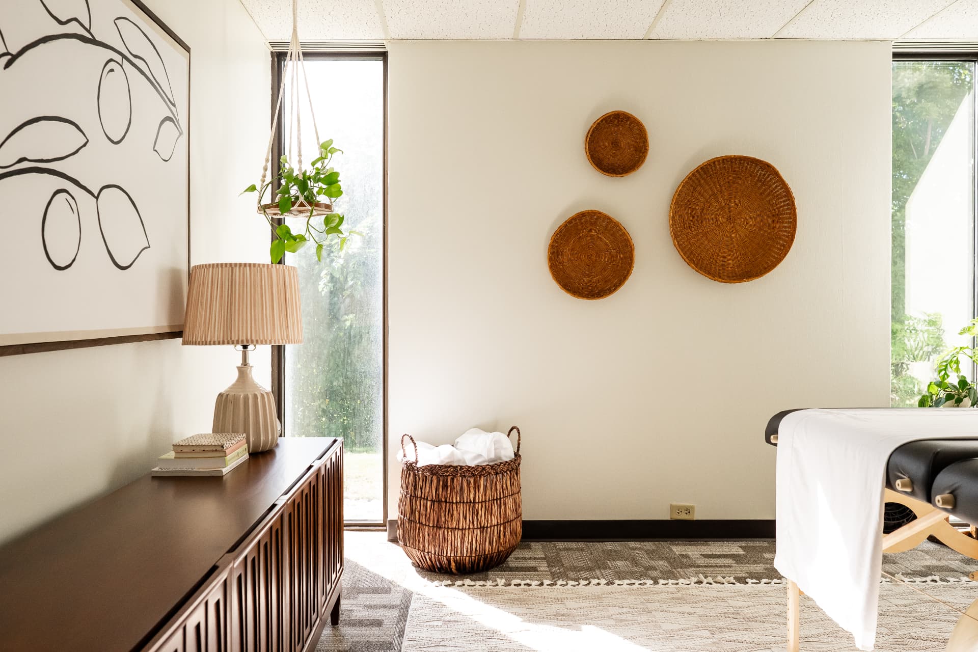 A sunlit treatment room with plants, woven decor, and a treatment table.